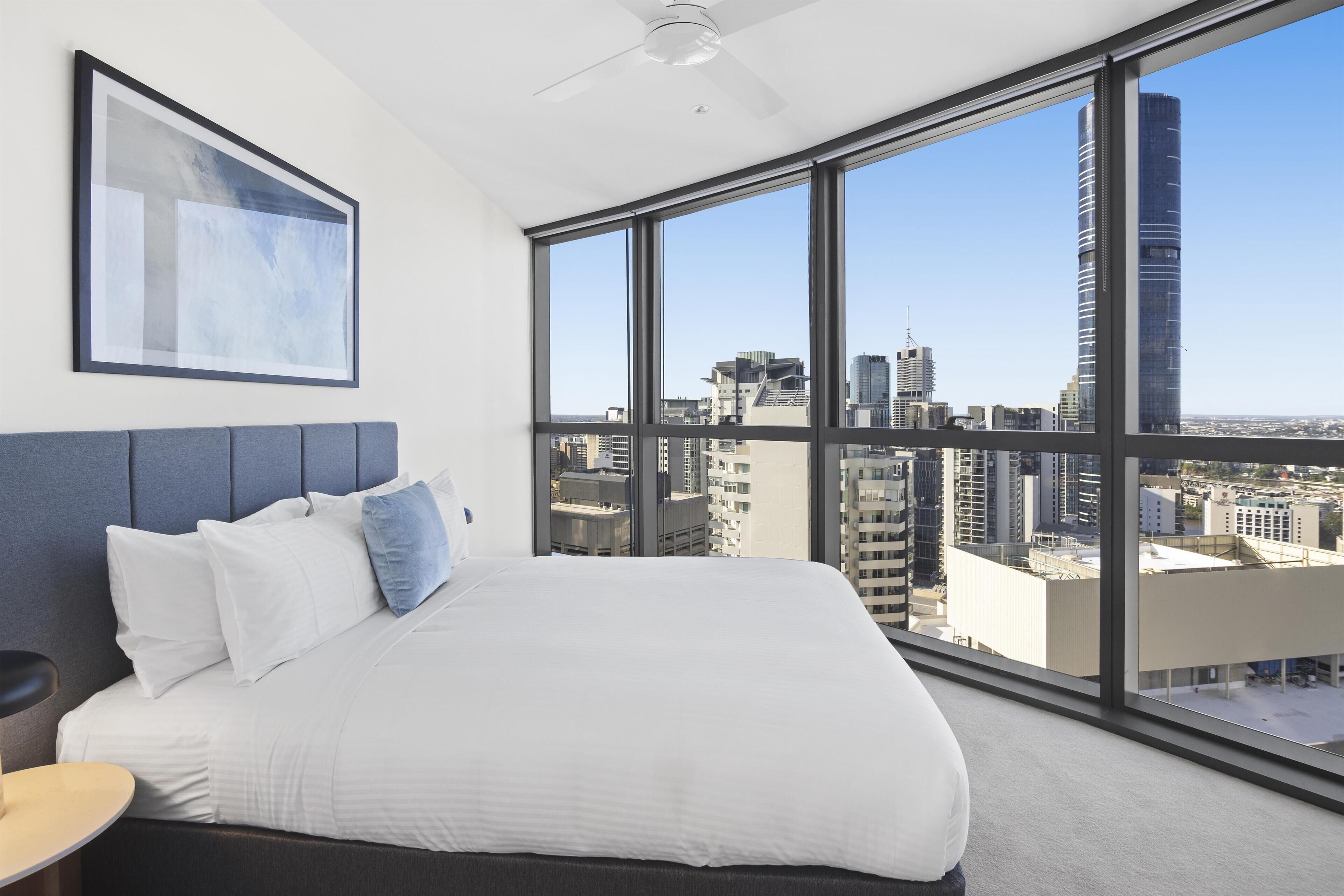 Ein Hotelzimmer mit einem großen Bett, einem Deckenventilator, einem gerahmten Kunstwerk und Blick auf die Stadt durch große Fenster.