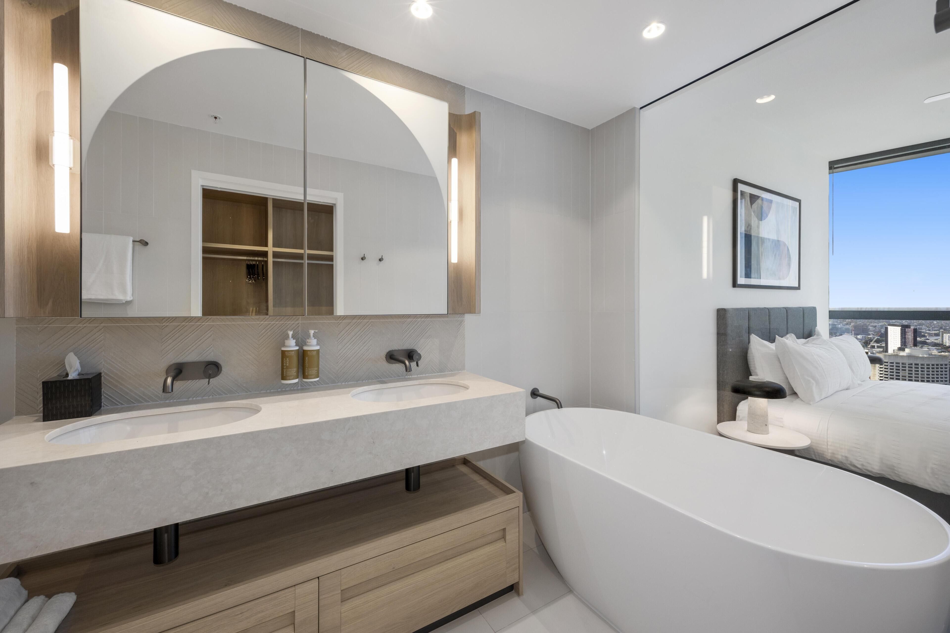 A modern bathroom with a large mirror, two sinks, a bathtub, and a city view.