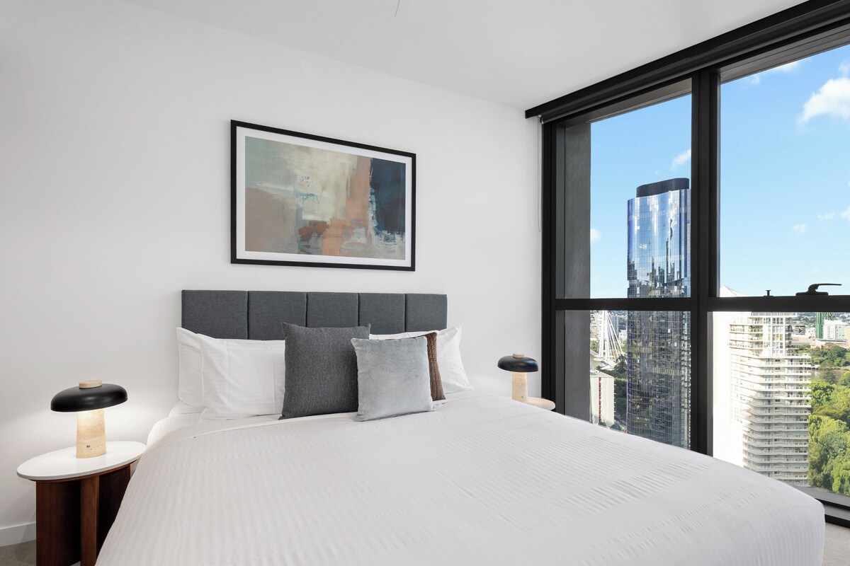 A modern hotel room with a large bed, a city view, and a framed abstract painting on the wall.