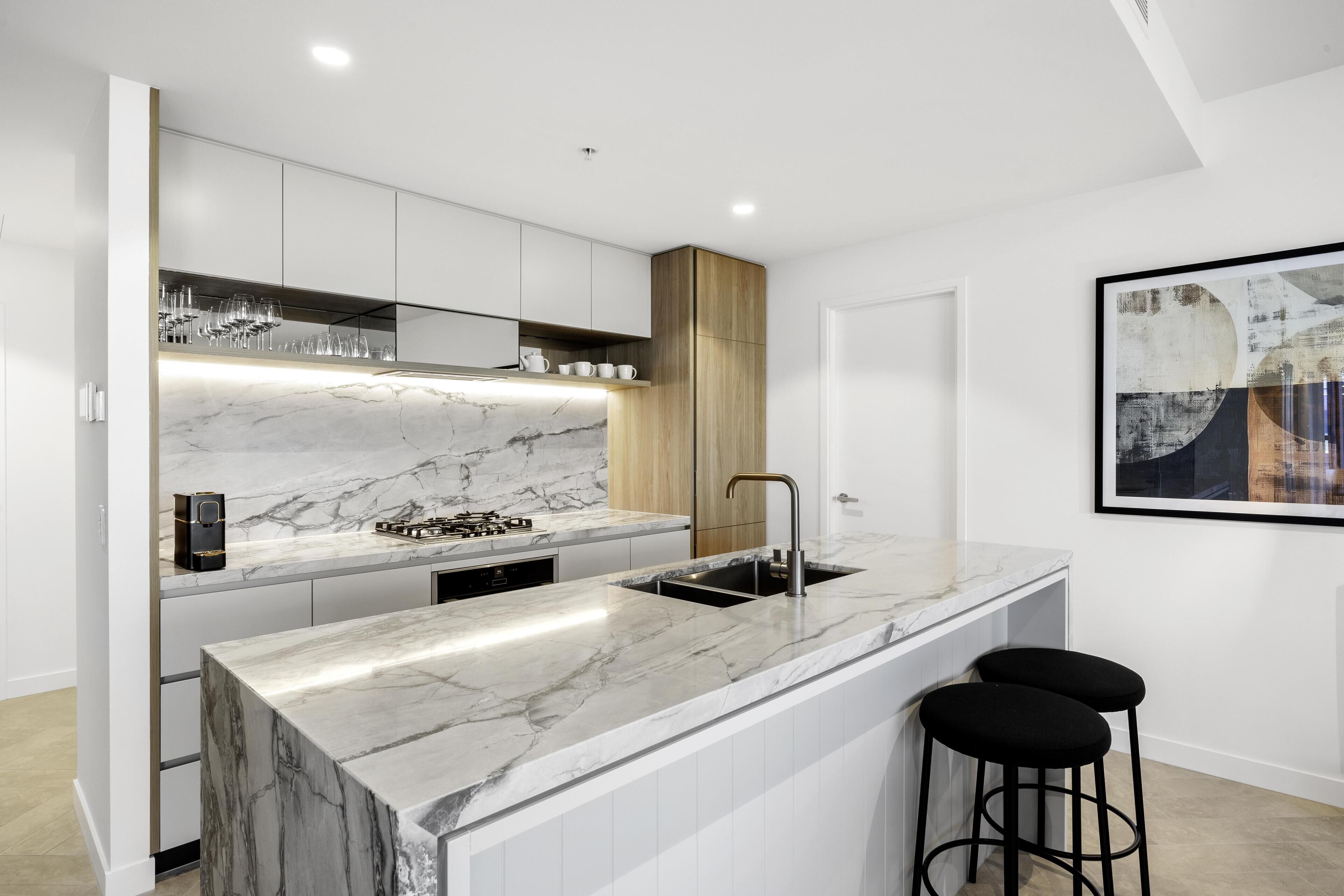 A modern kitchen with a marble countertop, wooden cabinets, a built-in sink, and two black bar stools.