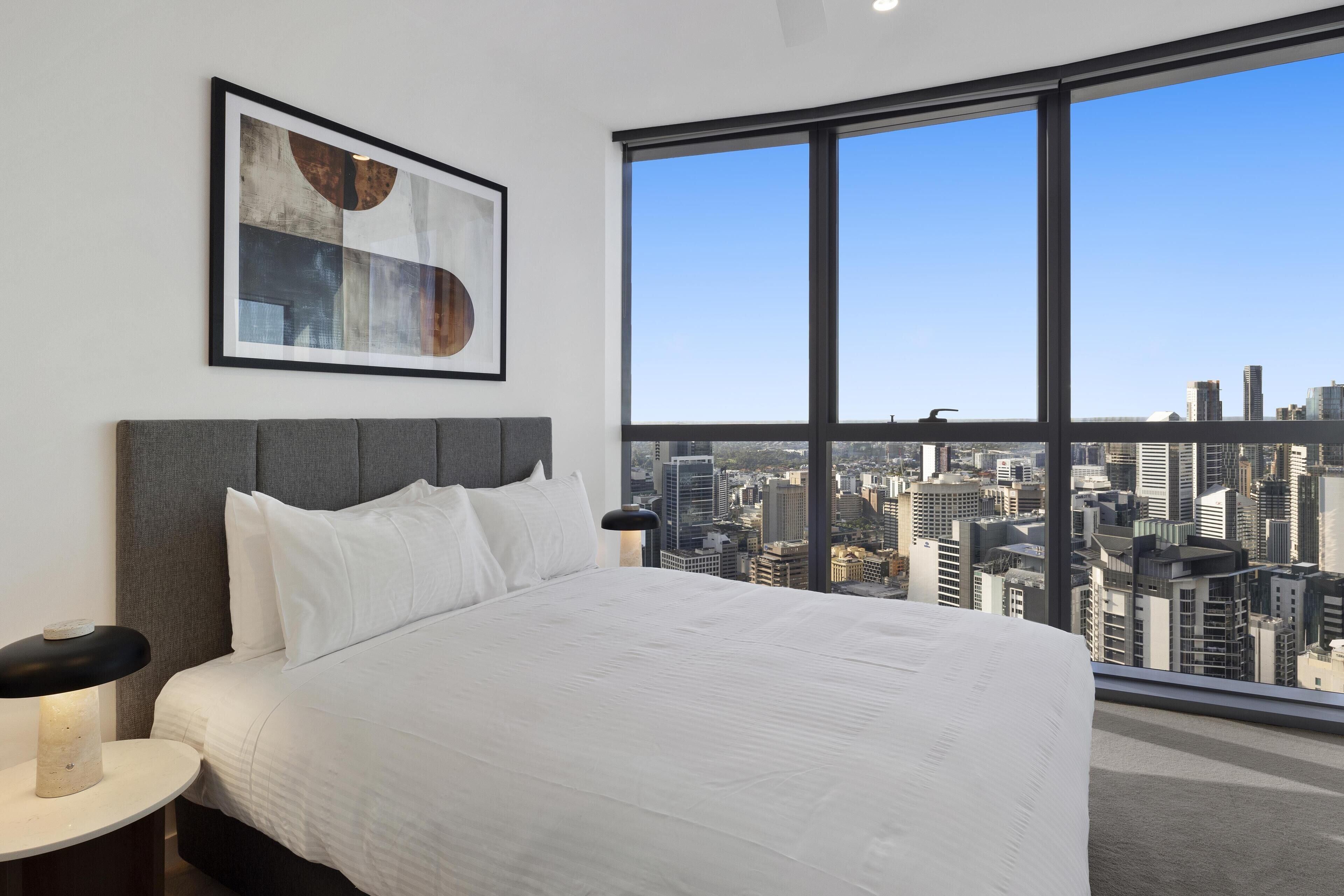 Quarto de hotel com uma cama grande, uma mesa de cabeceira, uma pintura abstrata emoldurada e vista para a cidade através de uma janela ampla.