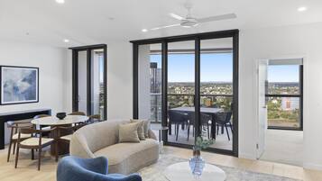 A modern living room with a sofa, chairs, a dining table, and a large window offering a city view.