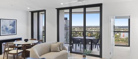A modern living room with a sofa, chairs, a dining table, and a large window offering a city view.