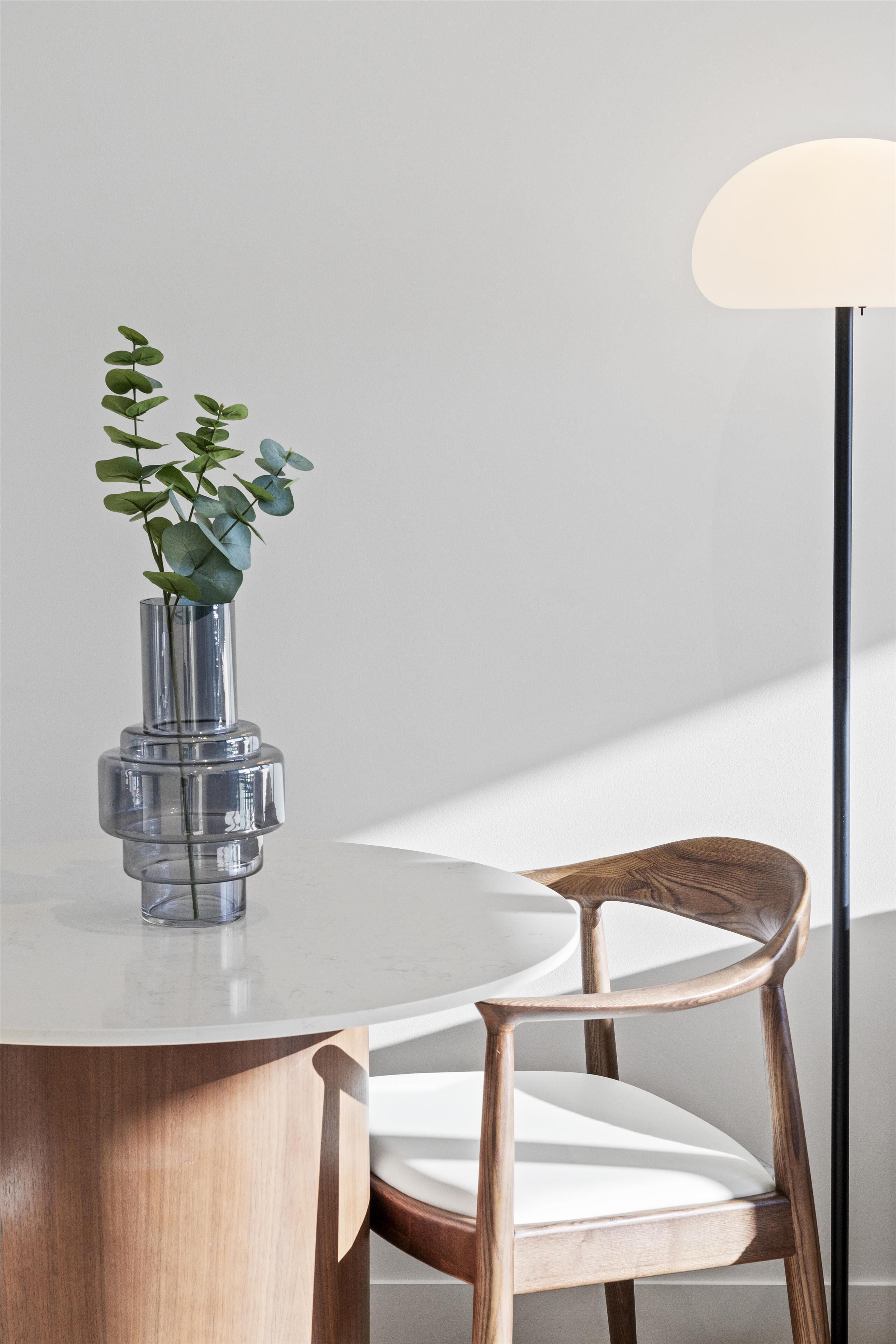 A round table with a clear glass vase containing green leaves, a wooden chair with a white cushion, and a floor lamp with a white shade.