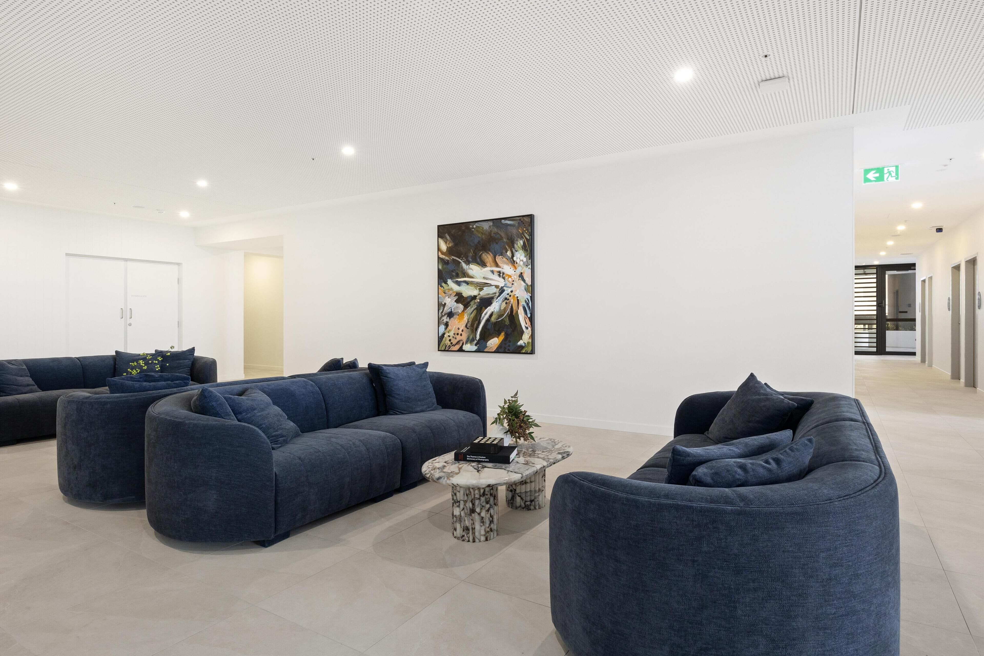 Uma área de recepção moderna com sofás azul escuro, uma mesa de centro de mármore e um quadro abstrato grande na parede.