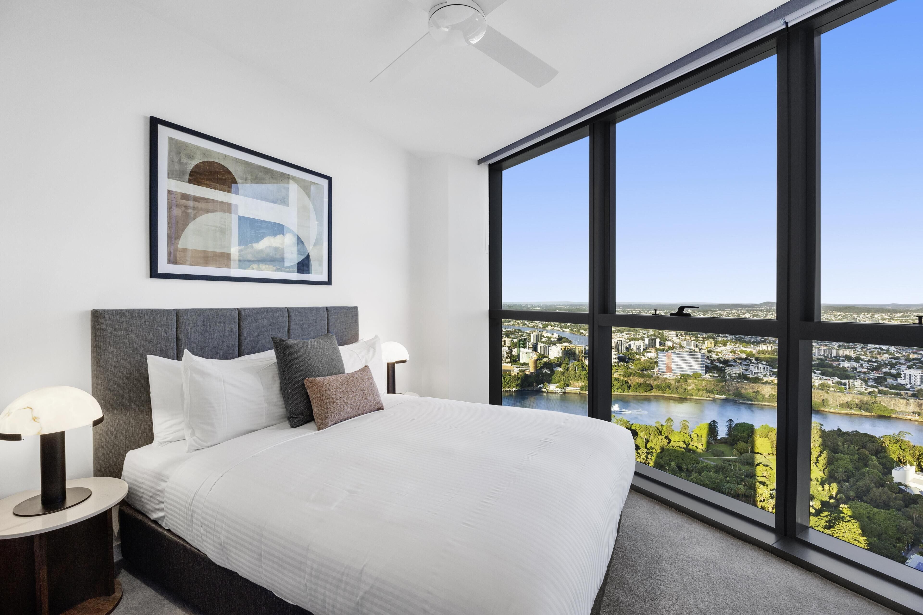 Une chambre d’hôtel moderne dotée d’un grand lit, d’un ventilateur de plafond et offrant une vue sur le paysage urbain et la rivière.