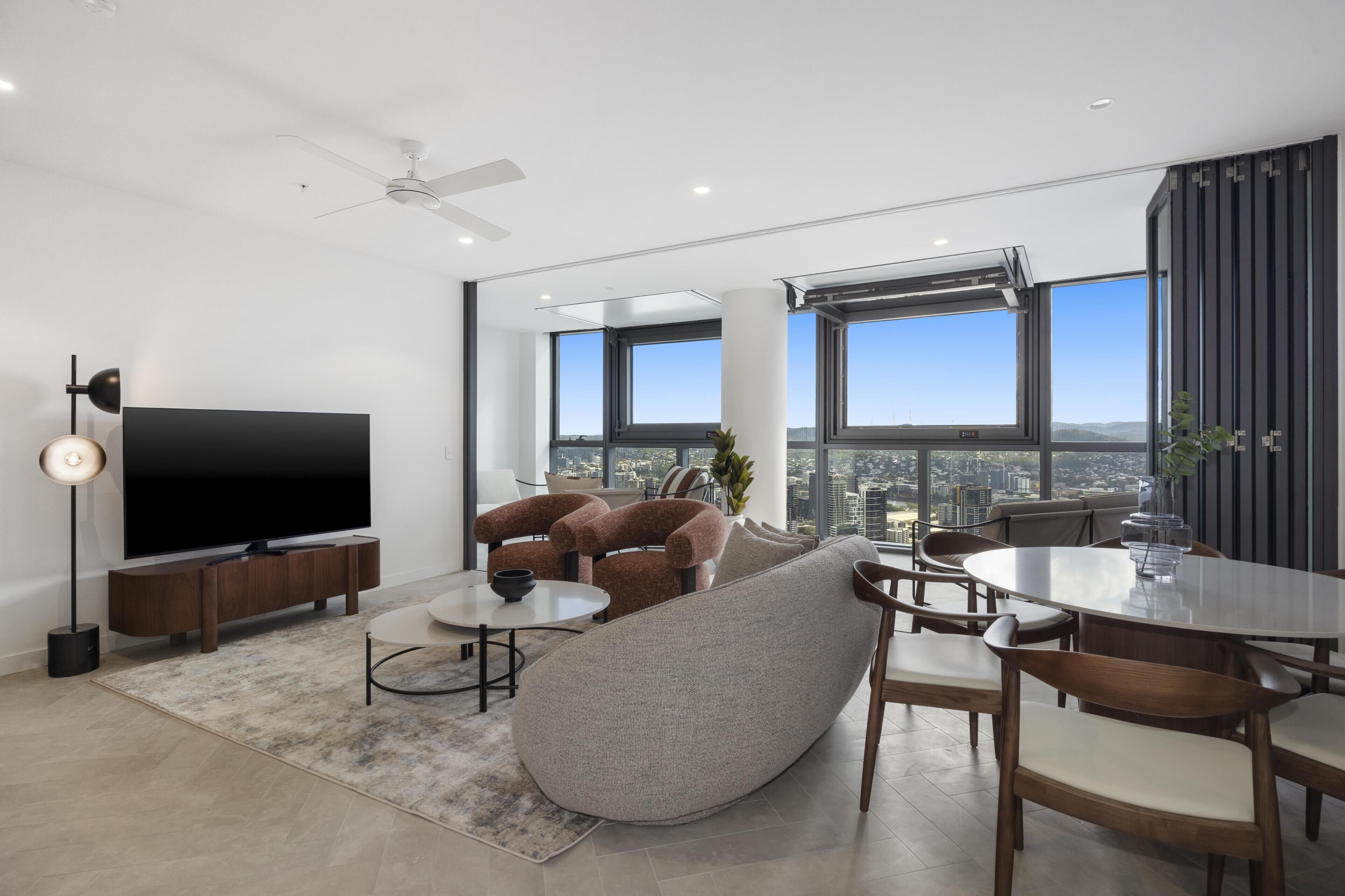 A modern living room with a large flat-screen TV, comfortable seating, a dining area, and large windows offering a city view.