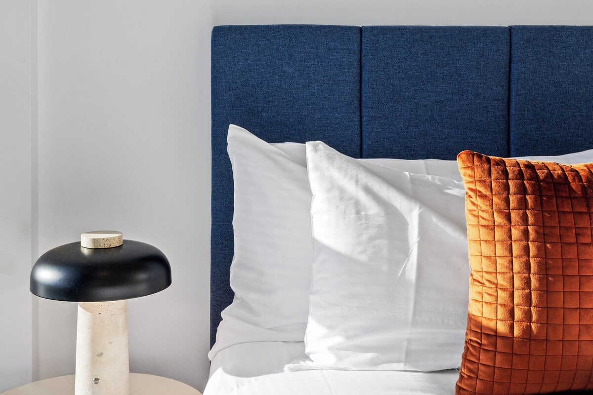 A bed with a blue headboard, white pillows, and a black and white lamp on a bedside table.