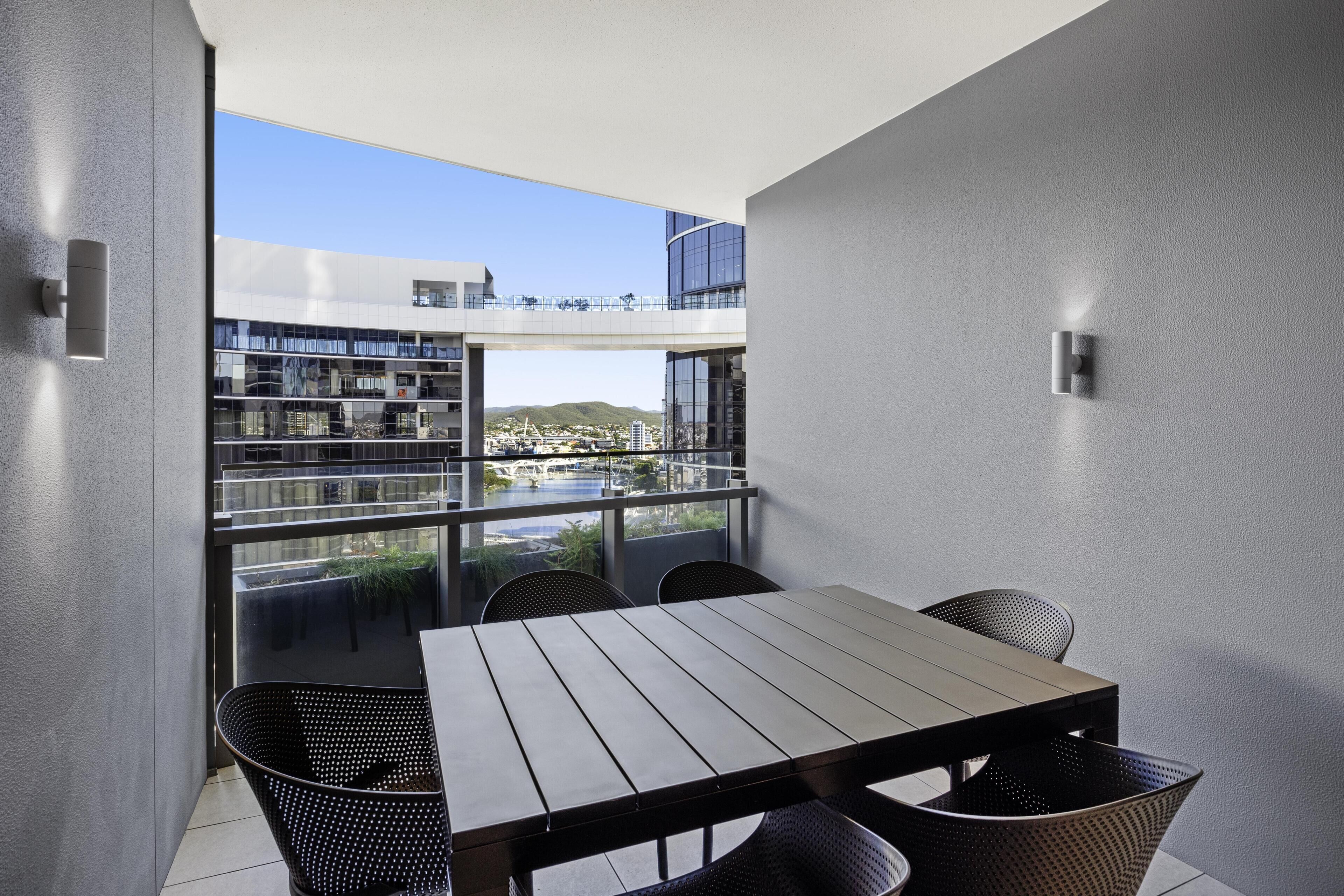 A modern balcony with a table and chairs, large windows offering a view of a cityscape and greenery.