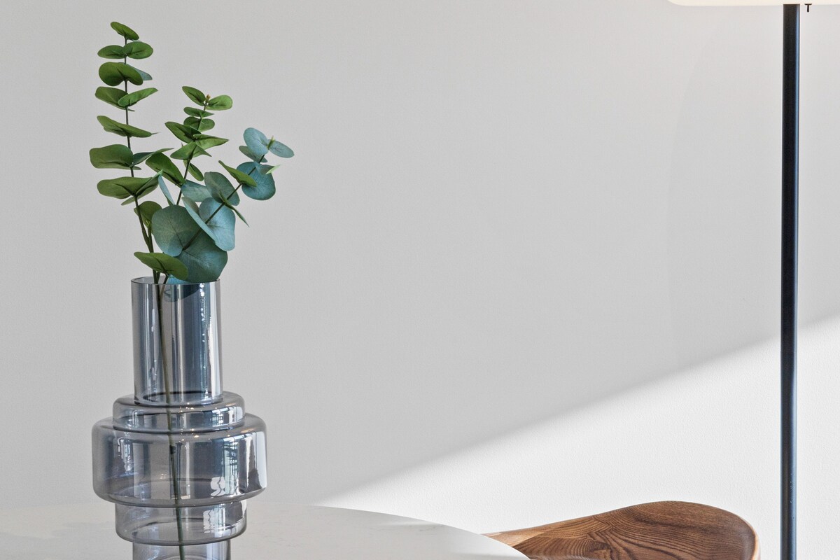 A round table with a clear glass vase containing green leaves, a wooden chair, and a white wall in the background.