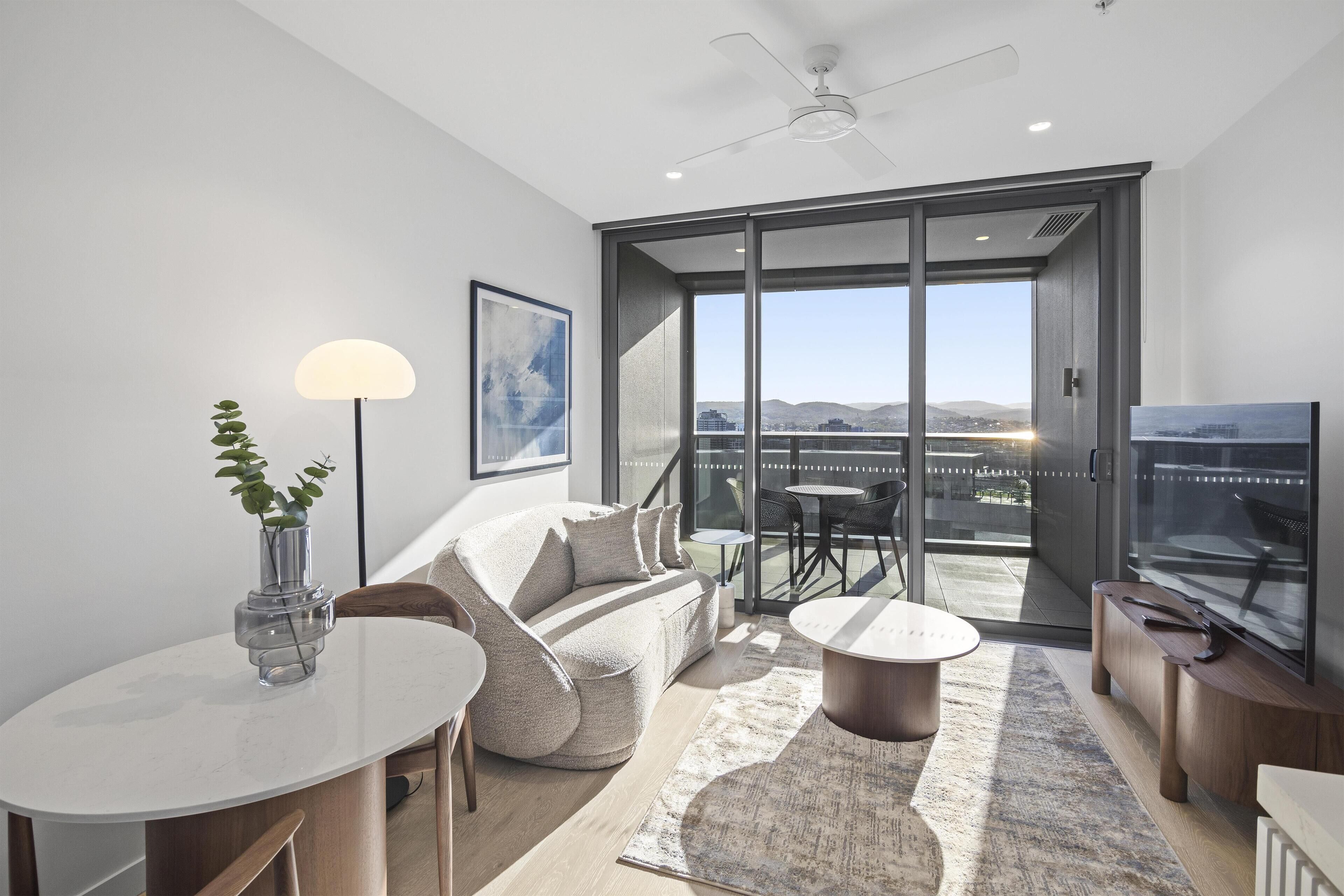 Una sala moderna con sofá, mesa de centro y televisor. Tiene un balcón con vista a la ciudad.
