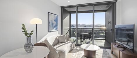 Una sala moderna con sofá, mesa de centro y televisor. Tiene un balcón con vista a la ciudad.