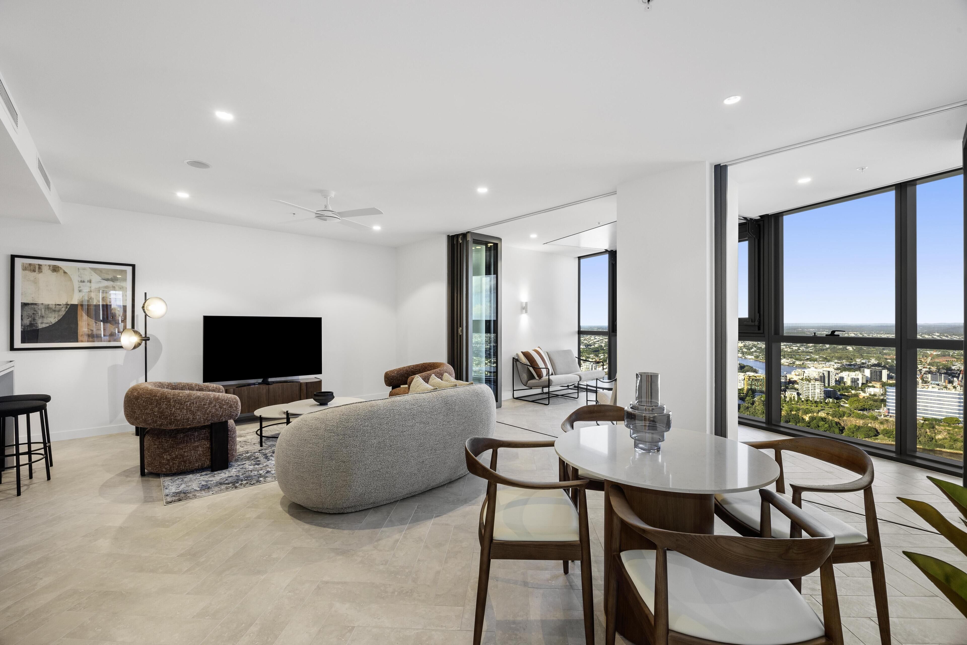A modern living room with a flat-screen TV, a sofa, a dining table with chairs, and large windows offering a city view.
