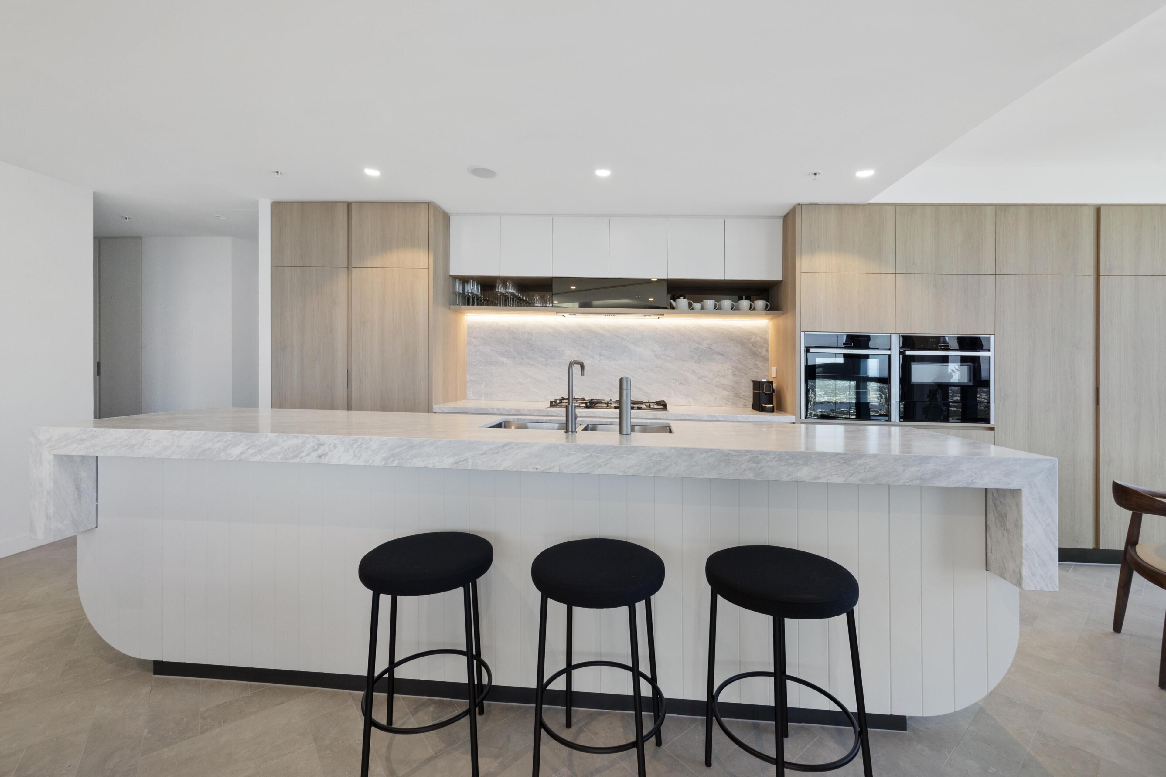 A modern kitchen with a marble countertop, three black bar stools, built-in appliances, and a city view.