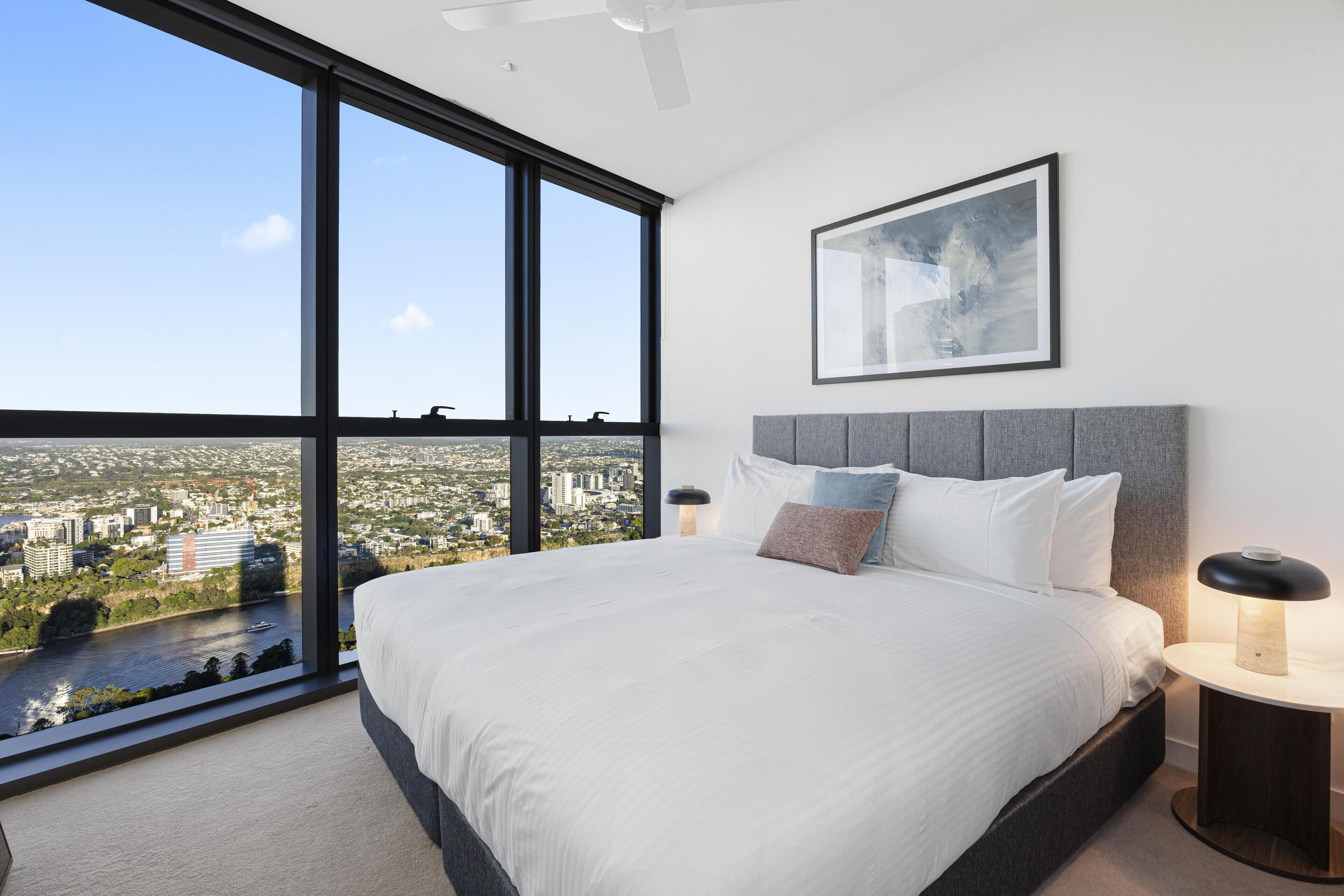 A modern bedroom with a large bed, a city view through floor-to-ceiling windows, a grey headboard, and a bedside table with a lamp.