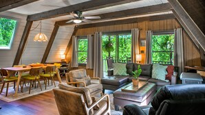 A spacious loft-style room with a dining area, seating, and large windows offering a view of greenery. - A-frame Cabin Near Mt. Mitchell | Rustic A-Frame on Private Acre (Burnsville)