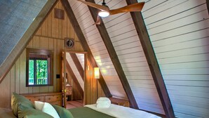 A cozy attic bedroom with a sloped ceiling, a wooden bed with pillows, a nightstand, and a window overlooking greenery. - A-frame Cabin Near Mt. Mitchell | Rustic A-Frame on Private Acre (Burnsville)