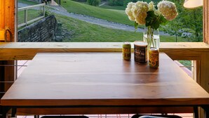 Uma mesa de madeira com um vaso de flores e duas latas de comida, com vista para uma paisagem deslumbrante com uma casa e montanhas.