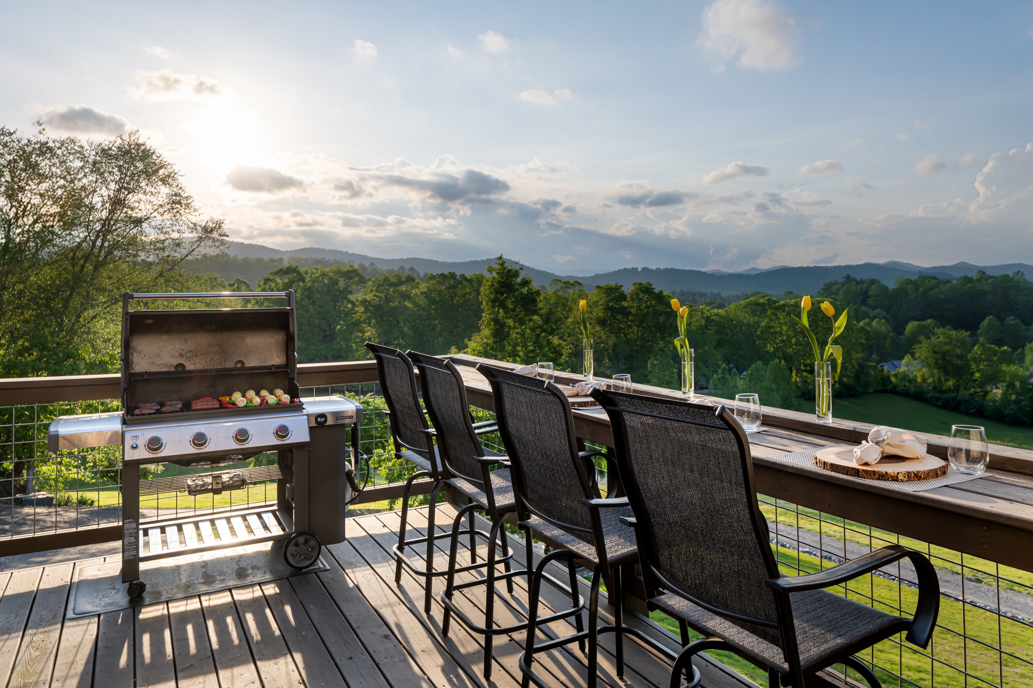A deck with a grill, chairs, and a scenic view of mountains and trees.
