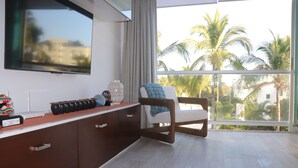 Een moderne woonkamer met een grote tv aan de muur, een houten stoel met een blauw kussen, en een uitzicht op palmbomen door grote ramen.