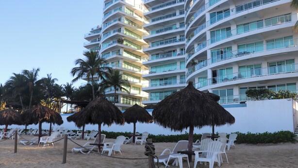 Een hotel aan het strand met meerdere verdiepingen, witte balkons en rieten parasols.