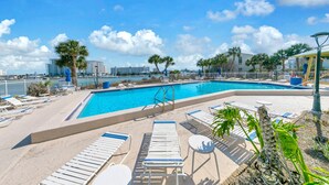 Piscina con sdraio e palme, circondata da una recinzione e con edifici sullo sfondo.