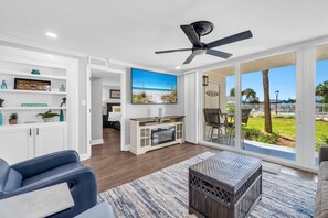 A modern living room with a flat-screen TV, a ceiling fan, and a view of a pool and palm trees.