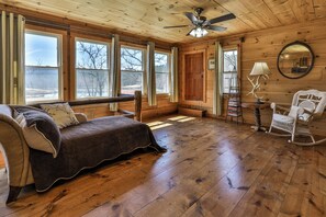 A cozy cabin interior with a wooden floor, a sofa, a rocking chair, a ceiling fan, and large windows. - NEW LISTING !!! RUSTIC MEADOW AT LADYBUG LAKE (Morganton)