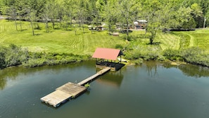 A lakeside wooden dock leading to a covered pavilion with a red roof, surrounded by lush greenery and a small house in the background. - NEW LISTING !!! RUSTIC MEADOW AT LADYBUG LAKE (Morganton)