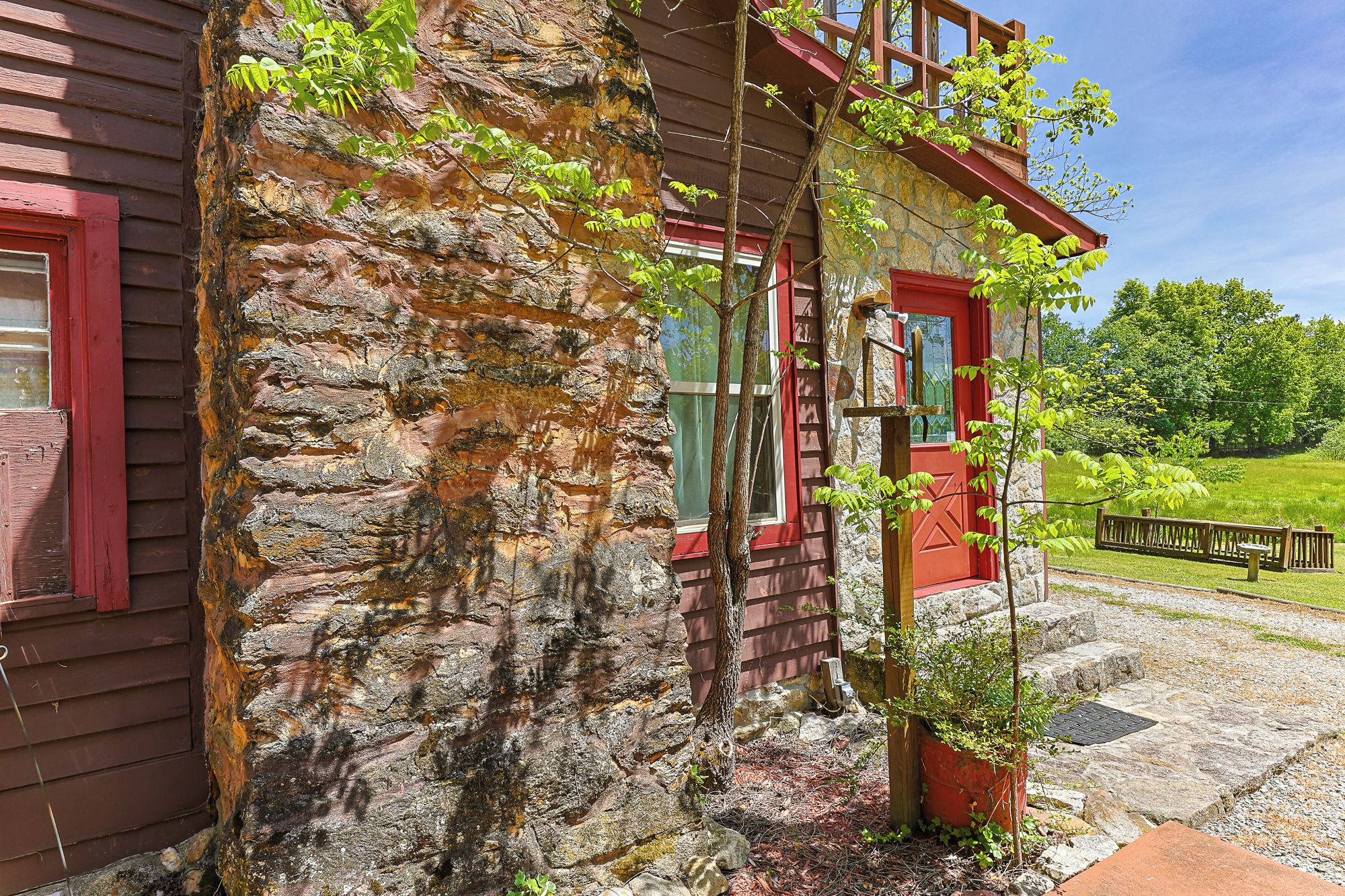 A rustic stone building with a red door and windows, surrounded by greenery and a wooden fence.