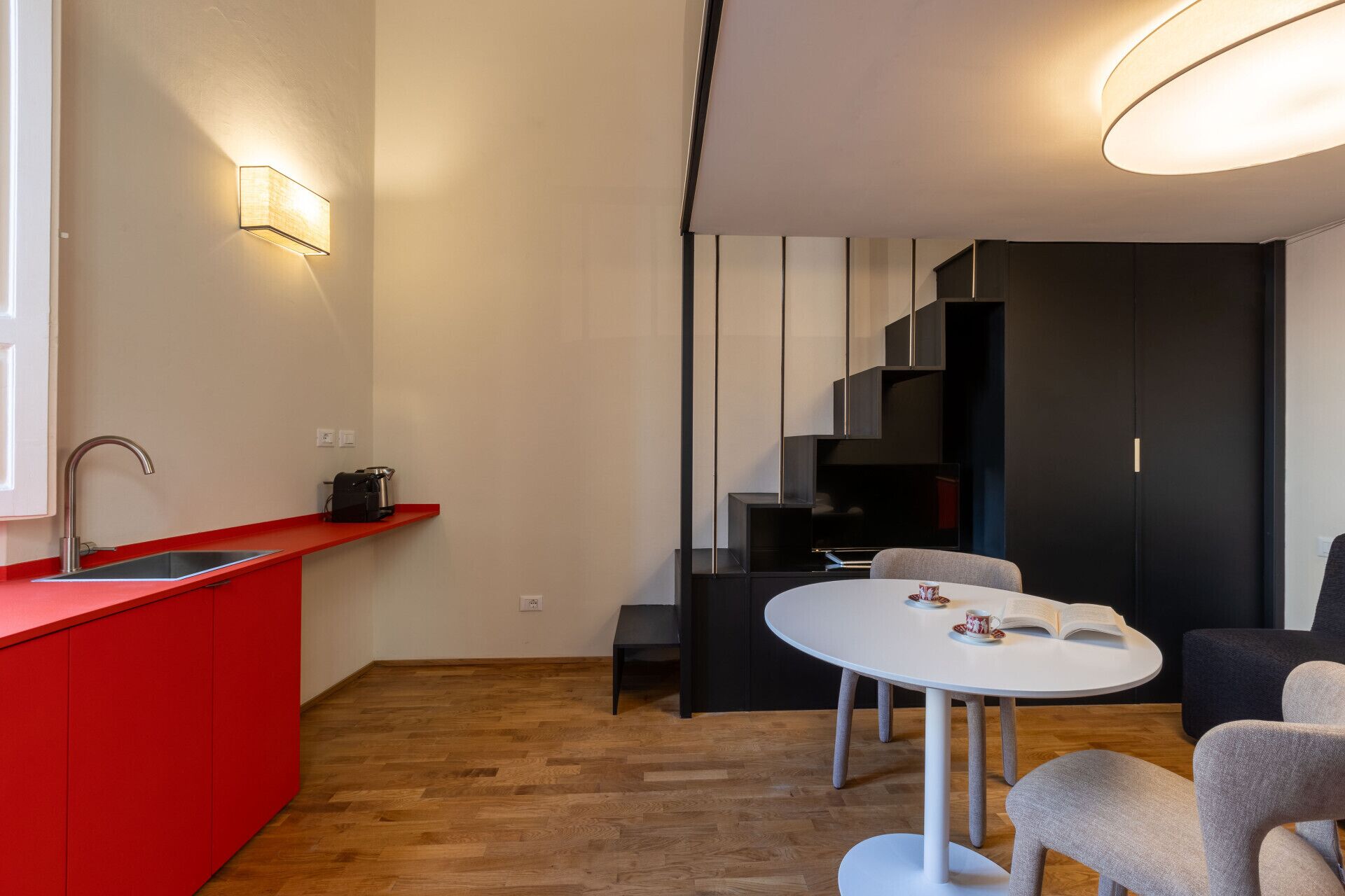 Una cocina moderna con encimera roja, un lavabo y una cafetera. Hay una zona de comedor con una mesa y sillas blancas.