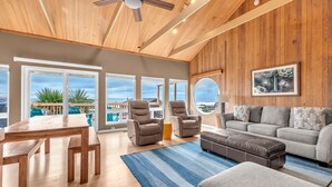 A spacious living room with a wooden ceiling, large windows, a gray sofa, leather chairs, and a dining area with a wooden table and chairs. - the Zephyr Shore in Roads End (Lincoln City)
