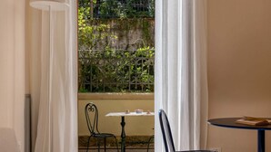 Ein Zimmer mit einem Bett, einem kleinen Tisch mit Stuhl und Blick auf einen Garten durch ein Fenster mit einem Metallgitter.