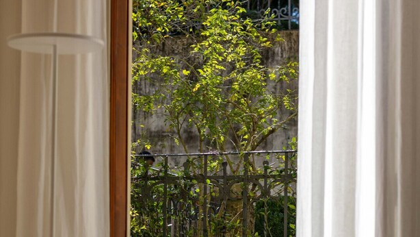 Ein Zimmer mit Blick auf einen Garten durch ein Fenster mit weißen Vorhängen. Es gibt einen Schachbrettboden, einen schwarzen Metallstuhl und einen Steinpflanztopf.