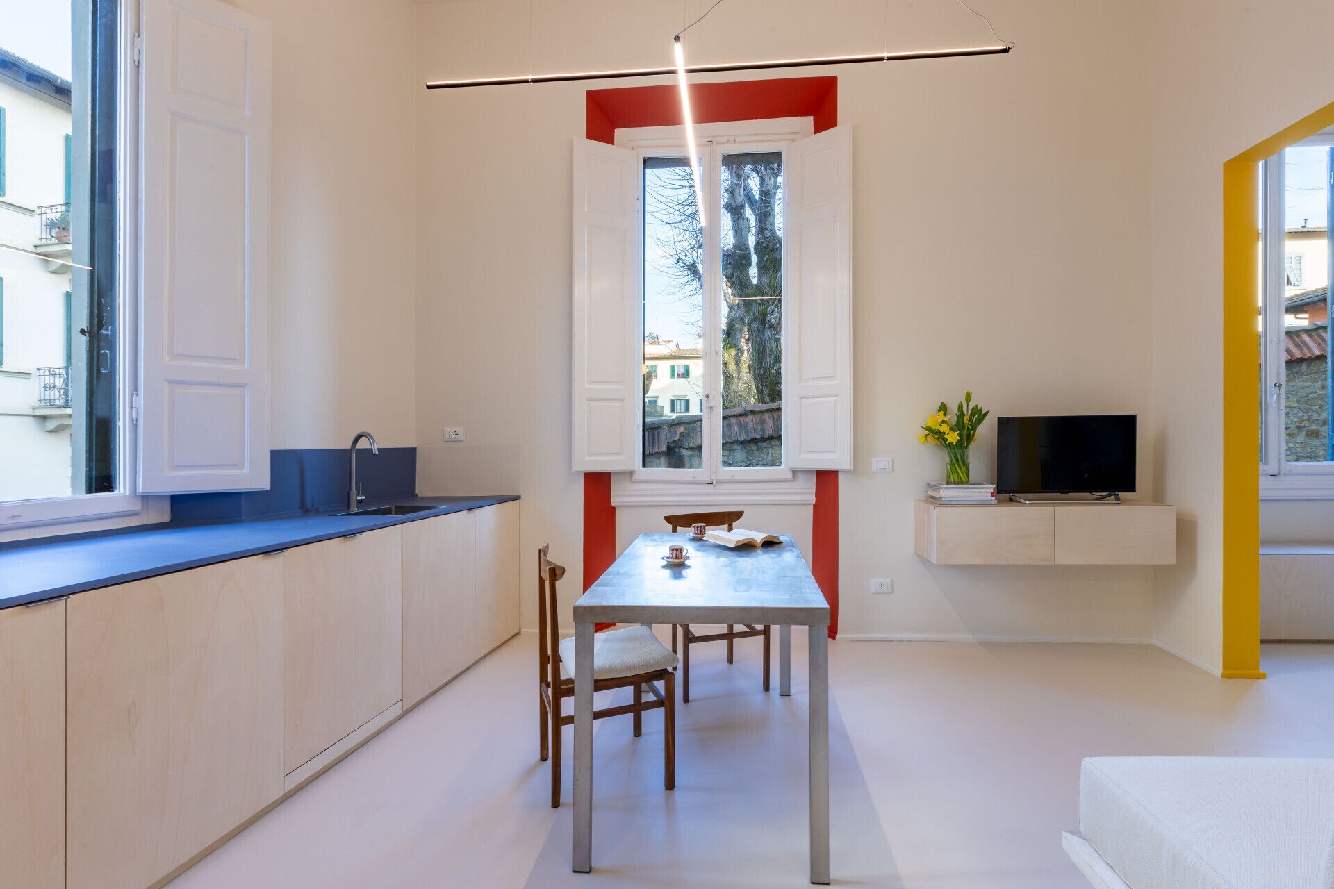 A compact kitchen with a dining area, a TV mounted on the wall, and a window with a view of buildings outside.