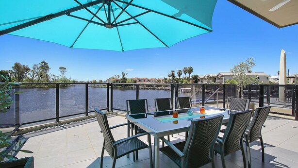 Un patio con mesa y sillas, con vista a un cuerpo de agua, y edificios y árboles al fondo.