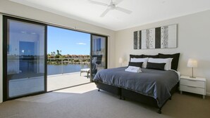 Un dormitorio moderno con una cama grande, ventilador de techo y vista a una masa de agua a través de amplios puertas corredizas.