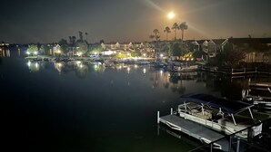 Una marina de noche con barcos amarrados, una hilera de casas con luces y una masa de agua tranquila que refleja las luces.