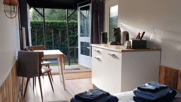 Quarto compacto com piso de madeira, uma mesa pequena com cadeira, cozinha compacta com cafeteira e vista para o verde através de uma porta de vidro.