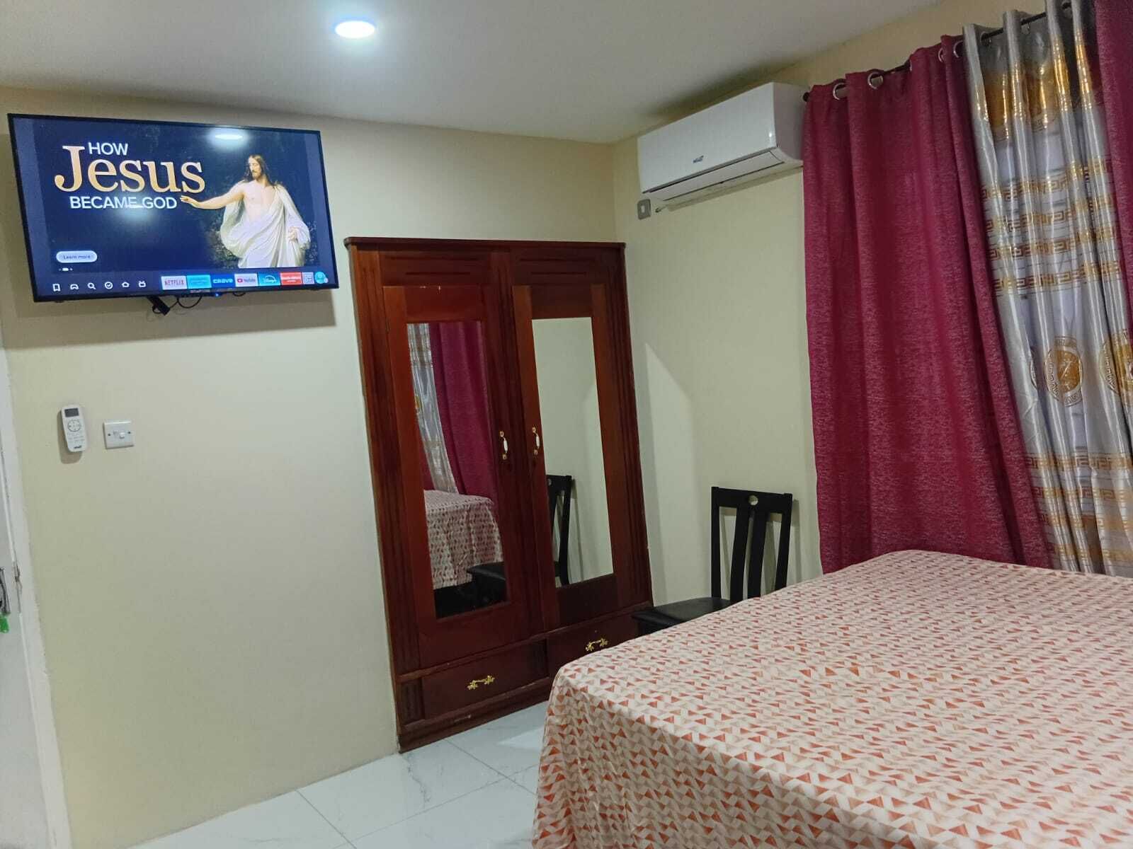 A hotel room with a bed, a wardrobe, a television displaying a program, and an air conditioning unit.