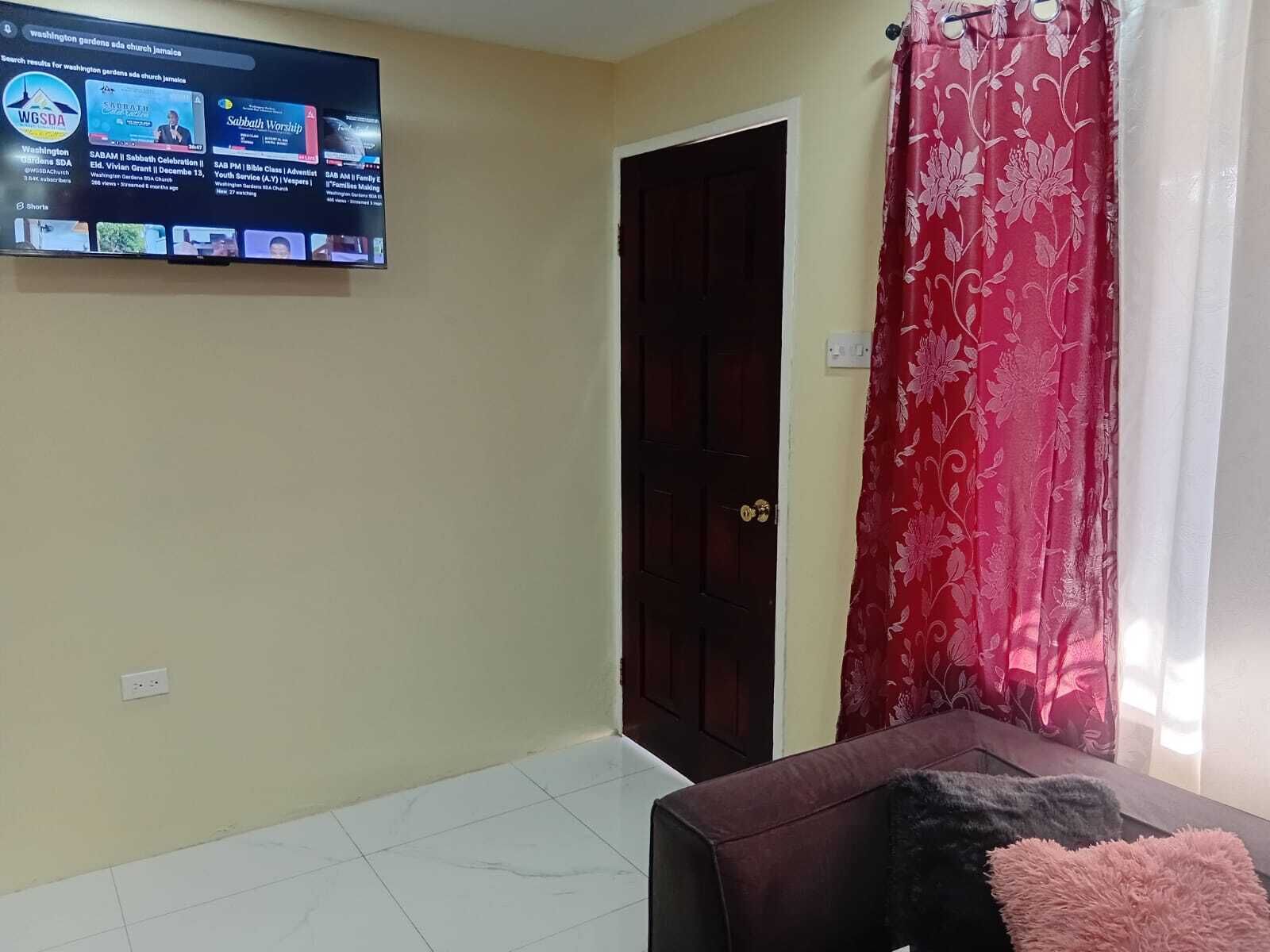 Ein Zimmer mit an der Wand befestigtem Fernseher, der einen Live-Stream einer Kirchenveranstaltung zeigt, einer geschlossenen Tür und einem Fenster mit rotem Blumenvorhang.
