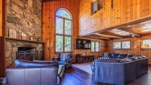 A log cabin interior with a stone fireplace, leather seating, and large windows. - Spacious Chalet Near Stratton and Mt. Snow w/Indoor Hot Tub, Fireplace, Firepit (Wardsboro)