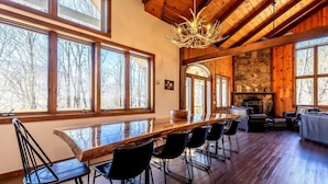 A spacious dining area with a long wooden table and black chairs, large windows with a view of trees, and a stone fireplace. - Spacious Chalet Near Stratton and Mt. Snow w/Indoor Hot Tub, Fireplace, Firepit (Wardsboro)