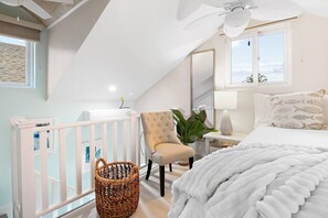 A bedroom with a white bed, a beige armchair, a white nightstand, a lamp, a wicker basket, and a ceiling fan. - Pineapple Villa -1 Bedroom + Sofa Bed (Nassau)