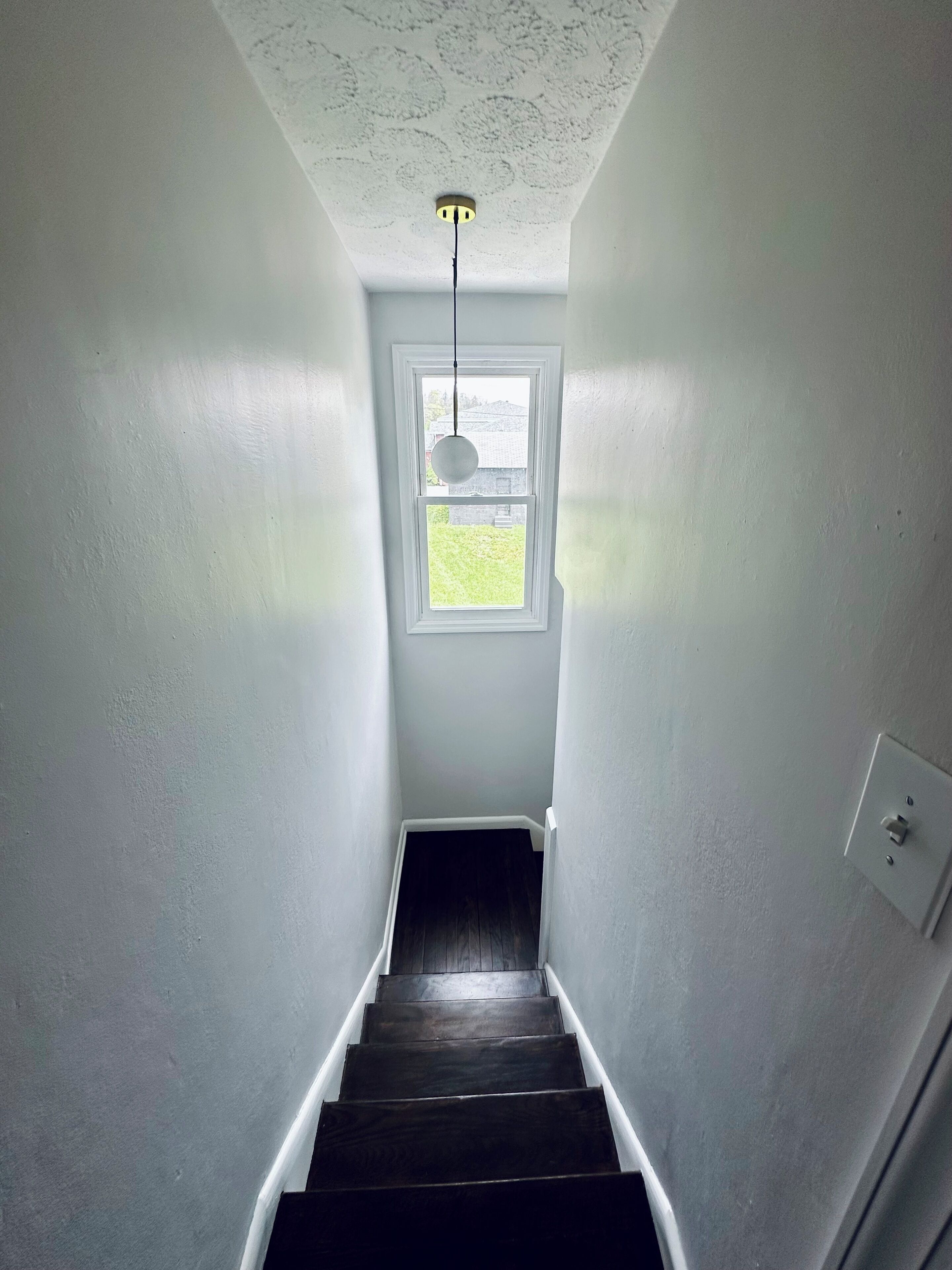 A narrow stairway with a window at the top, white walls, and a light fixture above.