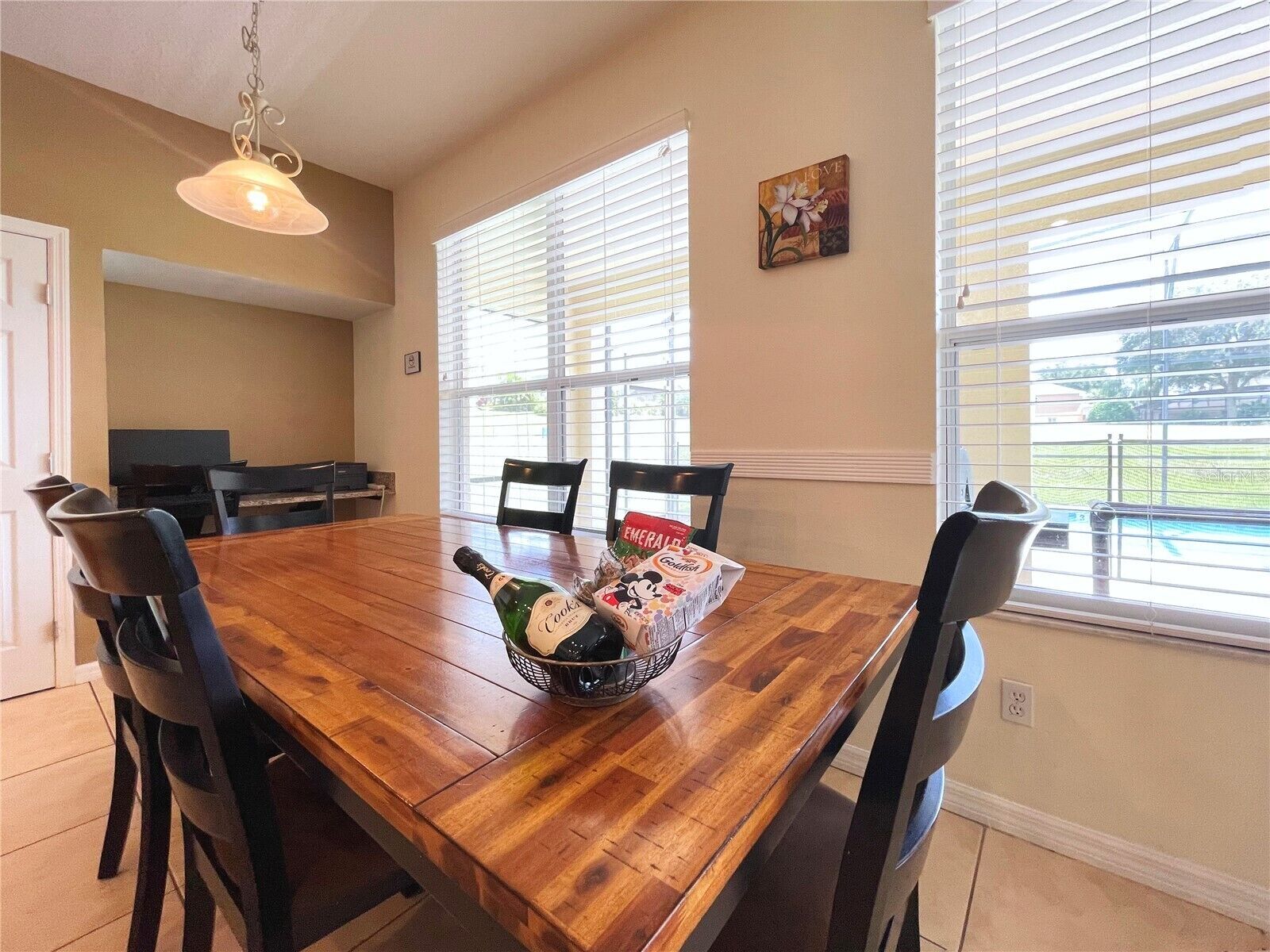 A dining area with a wooden table, chairs, a bottle of champagne, and a basket of snacks.