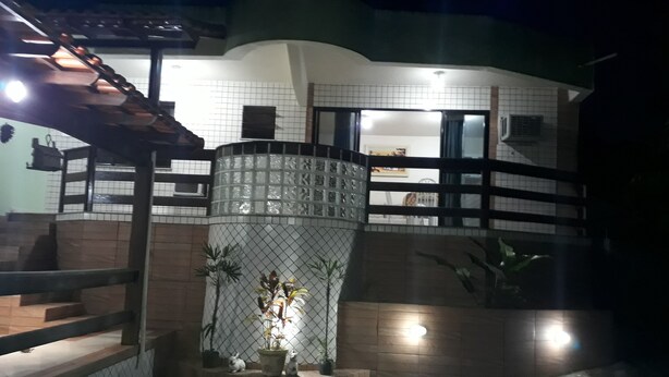 A residential building with a tiled exterior, a staircase leading to a balcony, and a fenced area with potted plants and a statue. - House in Guapimirim Condominium (Iconha Guapimirim)