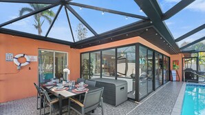 Un área de comedor al aire libre con piscina, rodeada por paredes de vidrio y una estructura de techo metálico.