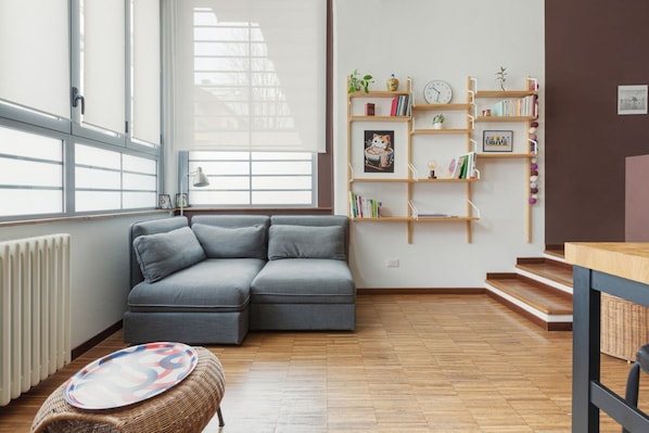 A modern living room with a grey sectional sofa, a wooden floor, a bookshelf, and a window with blinds.