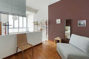 A room with a window, a chair, a sofa, a mirror, and a shelf with books and decorations.
